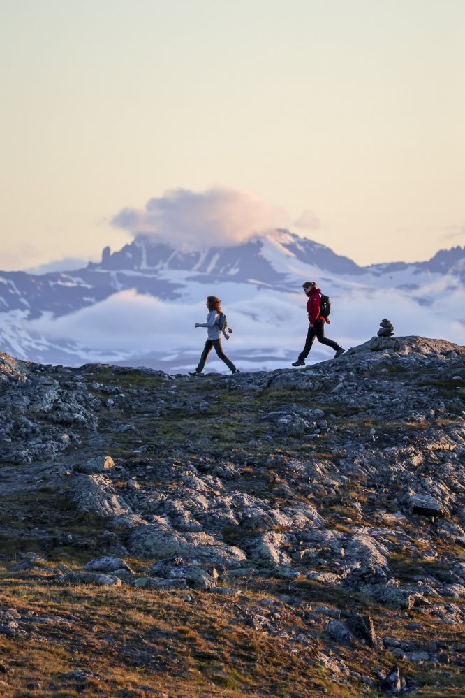 Kaksi naista kävelee sivuittain kameraan peräkkäin. Heidän takanaan on Norjan lumisia tuntureita, vaikka on kesäyö. Kuva on Saana-tunturin huipulta