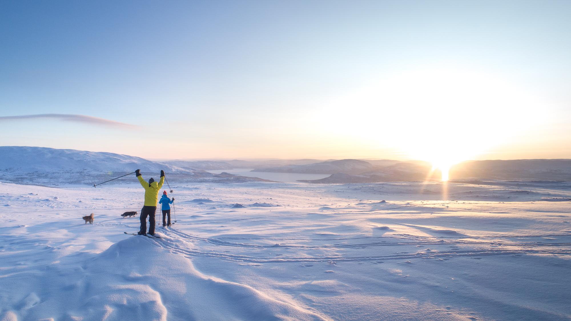 Two skiers and two dogs in Kilpisjärvi moving towards a beautiful sunset in February