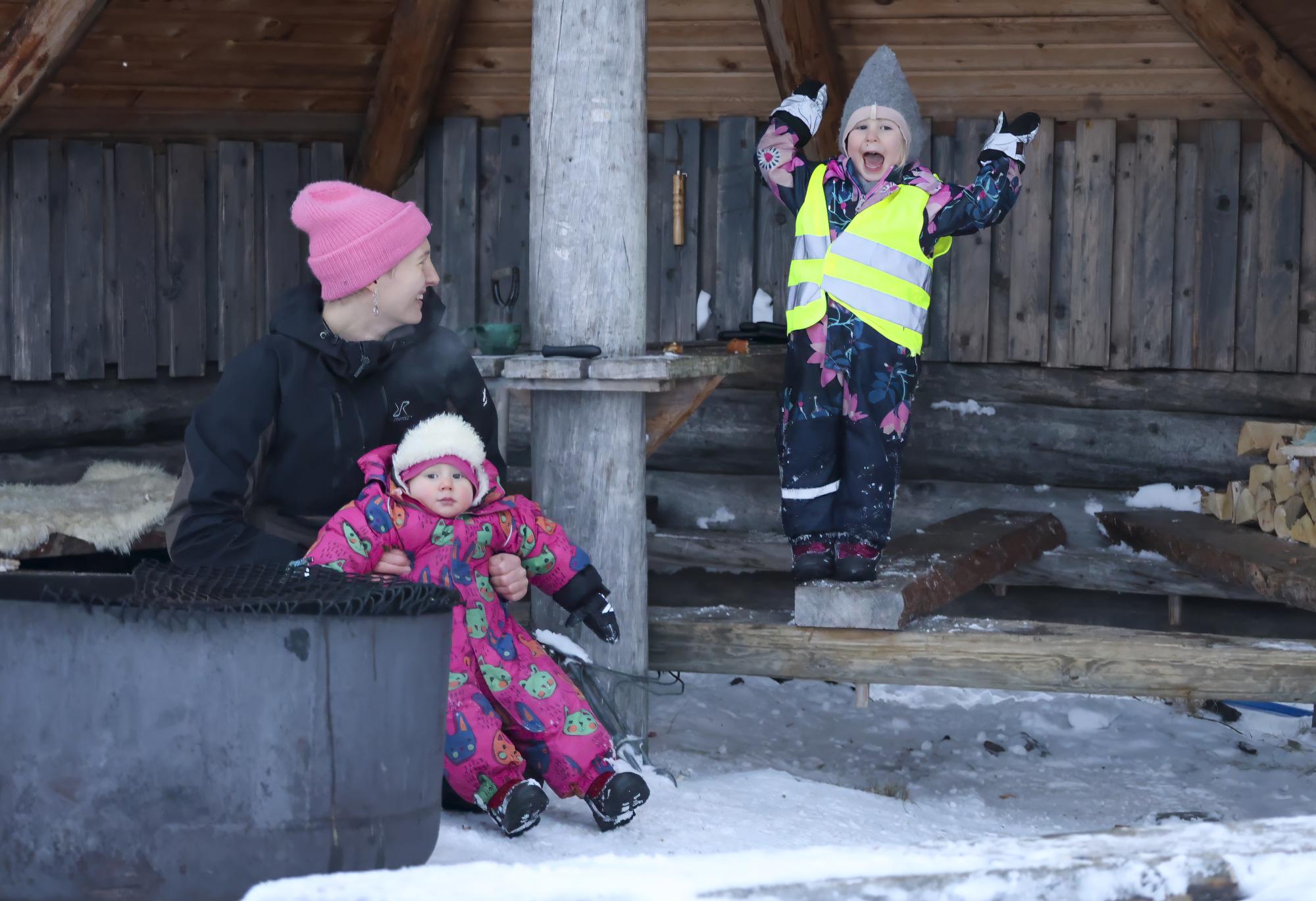 A mother and two children sit by the fire at the Tsahkaljärvi lean-to in winter. One of the children is smiling and raising their hands in joy.