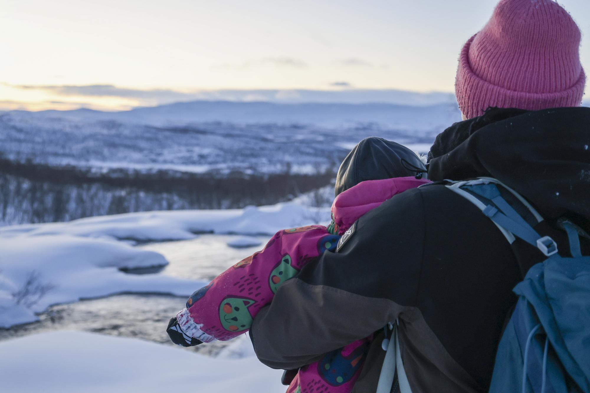 A mother in winter clothing, holding a child in her arms, stands with her back to the camera, looking out over the Tsahkal River and the landscape toward the village of Kilpisjärvi. The air is crisp with frost.
