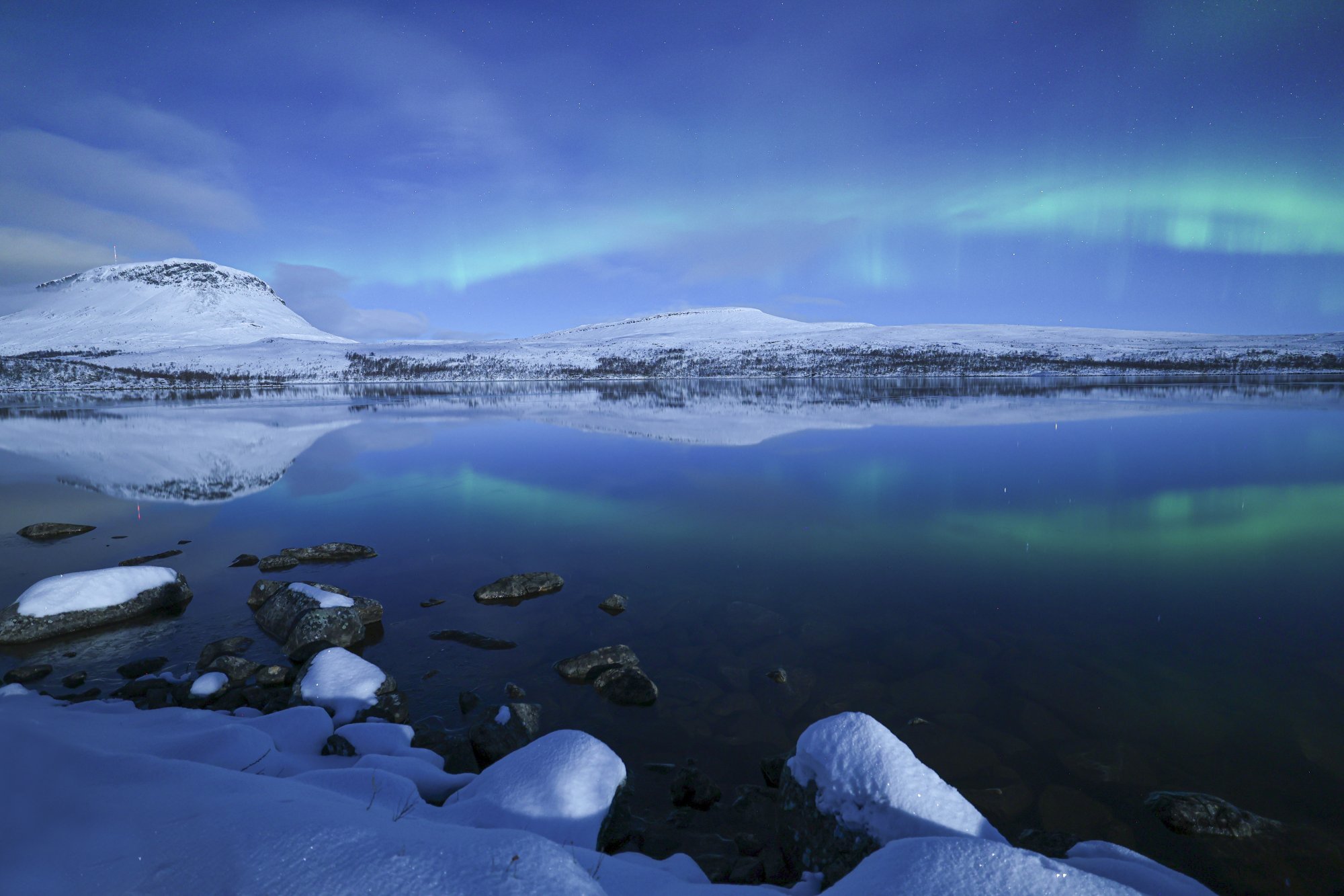 The Northern Lights are reflected on the surface of Tsahkaljärvi, with a snowy landscape and Saana fell visible in the moonlight in the background.