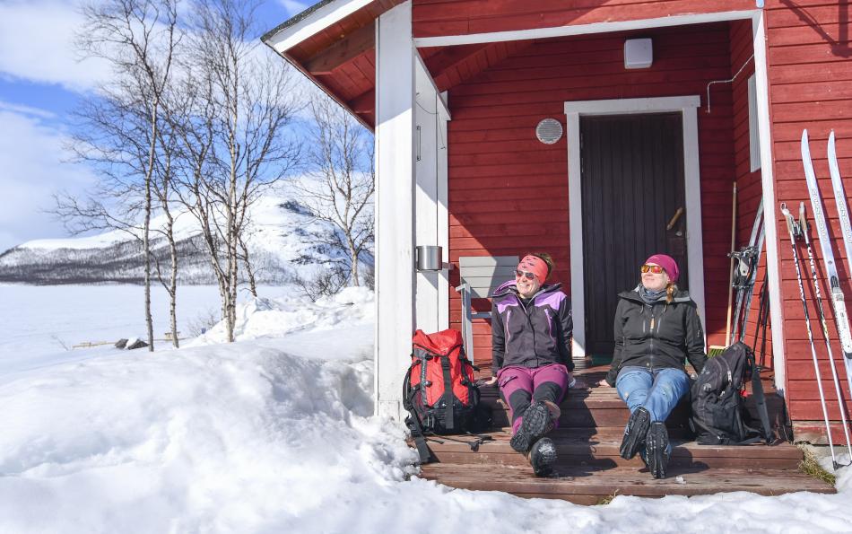 Two ladies with are sitting on a porch of a cabin at Kilpisjärven Retkeilykeskus. The sun is shining and they have just returned from a ski trip with their skis against the wall