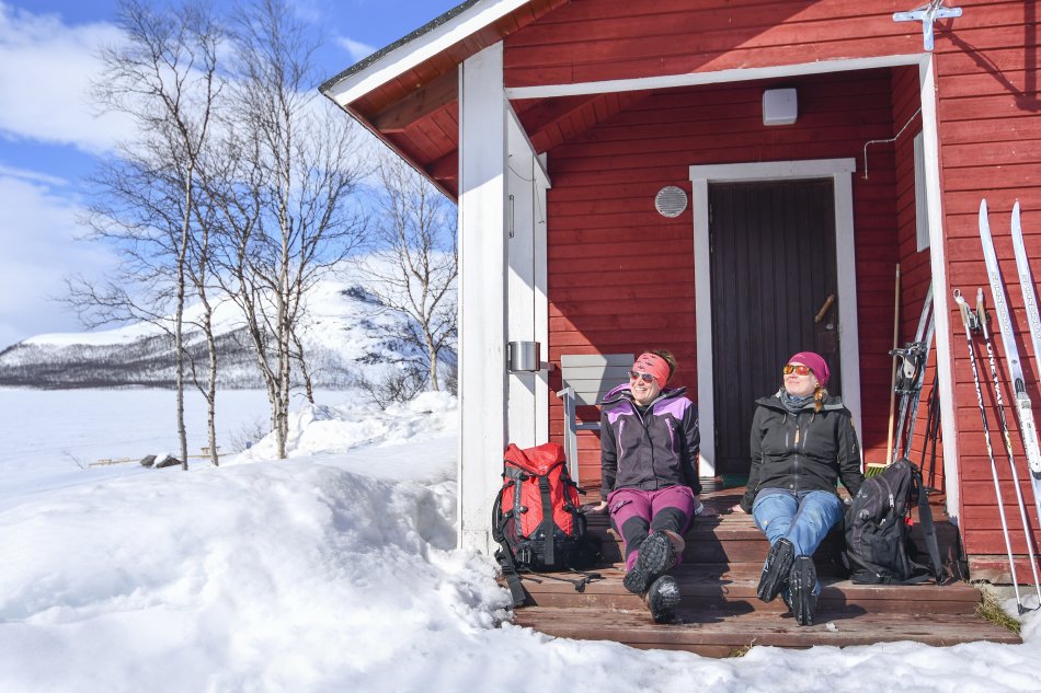 Two women are sitting in the sun on the steps of a small red cabin. They have their backbags and skis and poles out as well