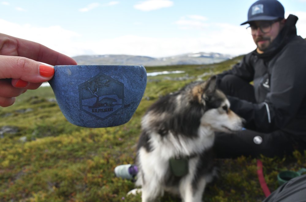 Kilpisjärvi-Kupilka uudessa mustikan värissä Kuvassa on naisen pitelemä Kilpisjärvi aiheinen muki taustalla tuntureita sekä mies ja koira tauolla