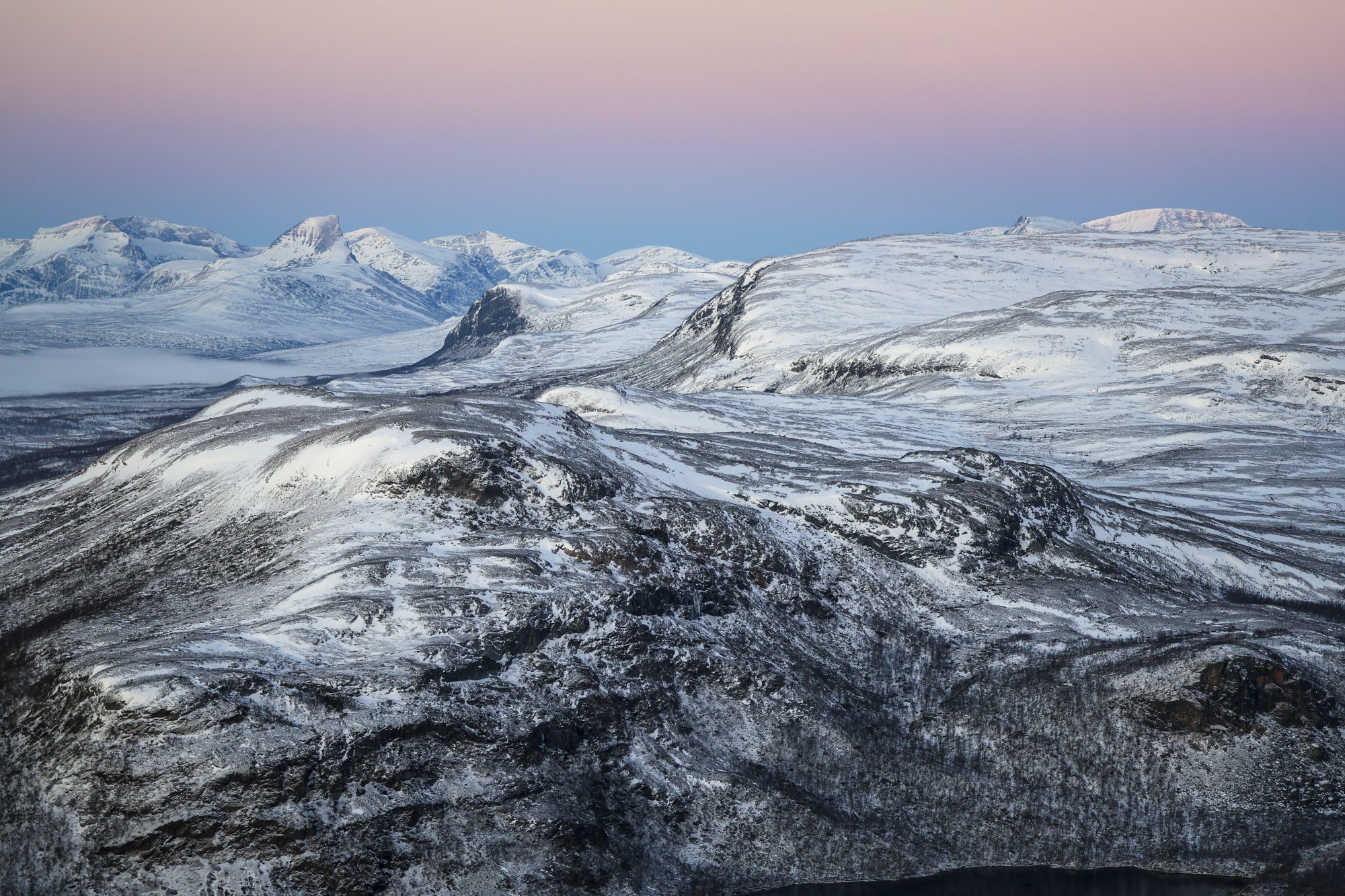 Maisemaa Saanalta kohti Norjaa marraskuussa. Lunta on tunturimaisemassa vähän, mustat kalliot ja jyrkänteet erottuvat selvästi. Taivas on vaaleanpunainen ja sininen.