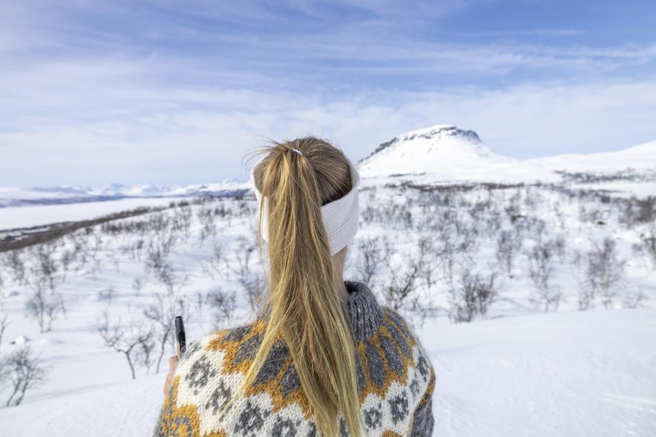 Women in colourful locally handmade sweater is standing outside in a wintery landscape looking at Saana fell.