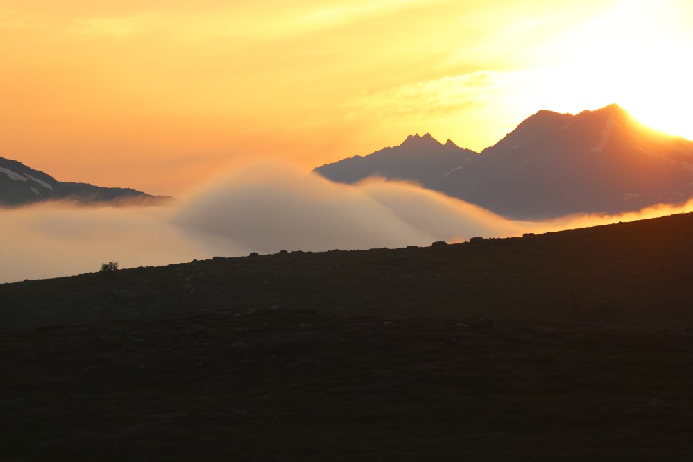 Aurinko nousee Norjan kahden vuorenhuipun takaa. Taivas on kirkkaan oranssi ja sumu nousee.