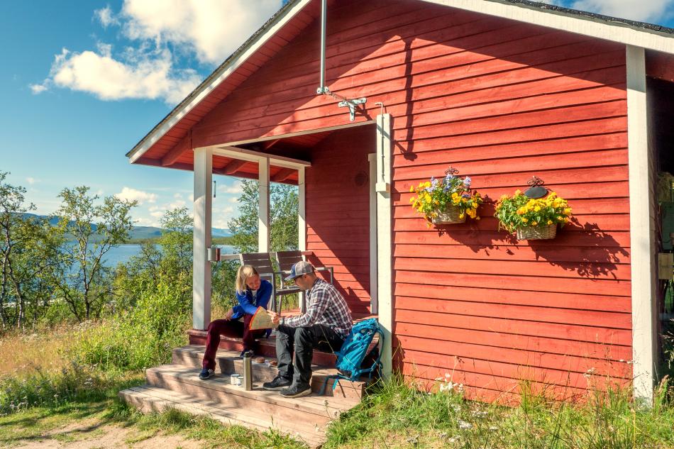 man and women are sitting on a steps of small red cabin in kilpisjärvi lapland finland. The sun is shining and the grass is green. There are colourful flowers on the cabin wall.
