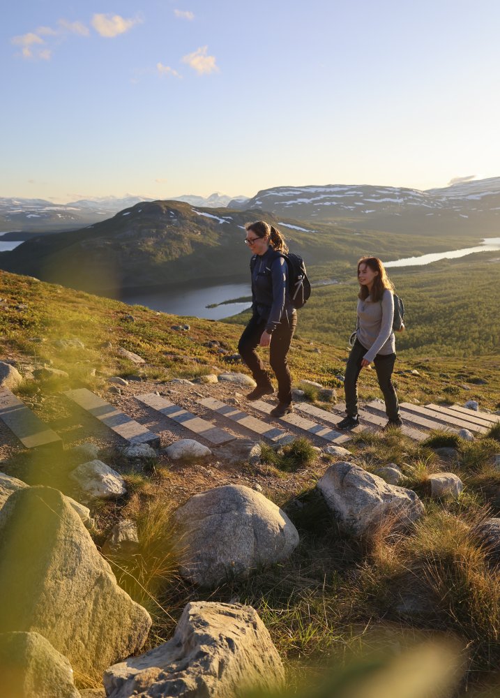 Kaksi naista kävelee Saanan portaita ylös kesäillan auringonpaisteessa. Taustalla näkyy Kilpisjärvi sekä pikku-Malla tunturi