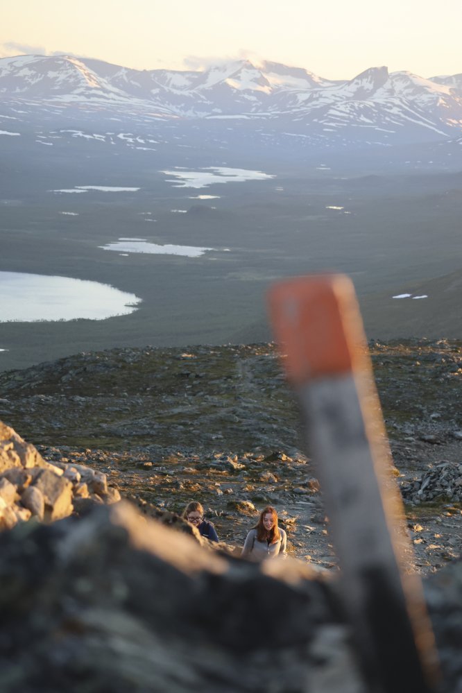 Reittimerkki Saanalla kesäyössä. Taustalla näkyy Norjan lumisia tuntureita sekä kaksi naista kiipeämässä huipulle.