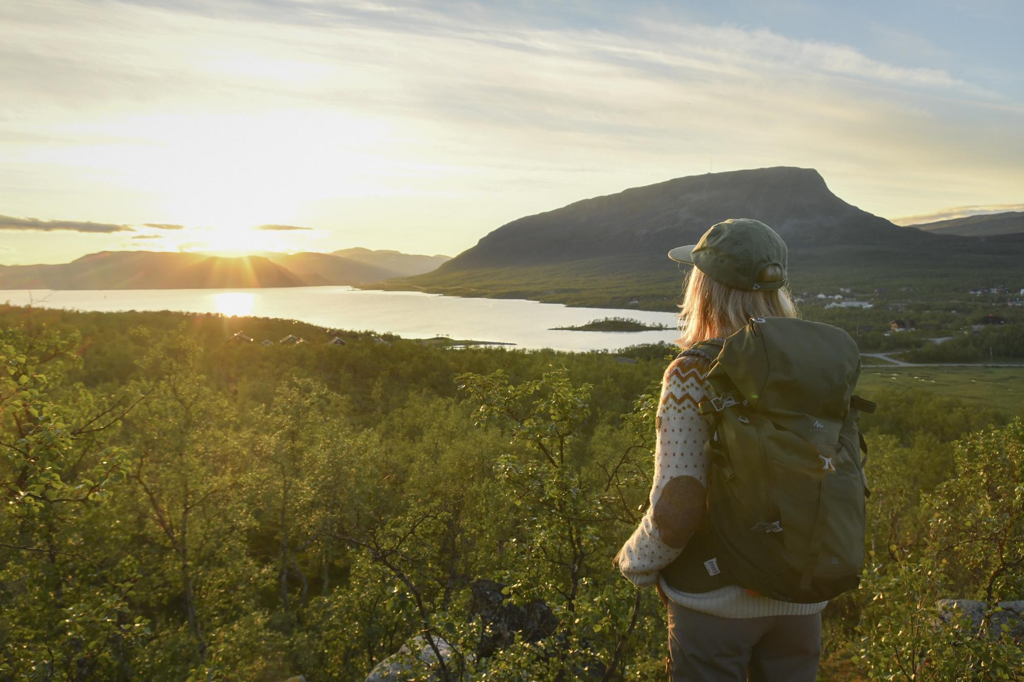 A woman with a backpack stands on the slopes of Salmivaara in Kilpisjärvi, gazing at the midnight sun of summer.