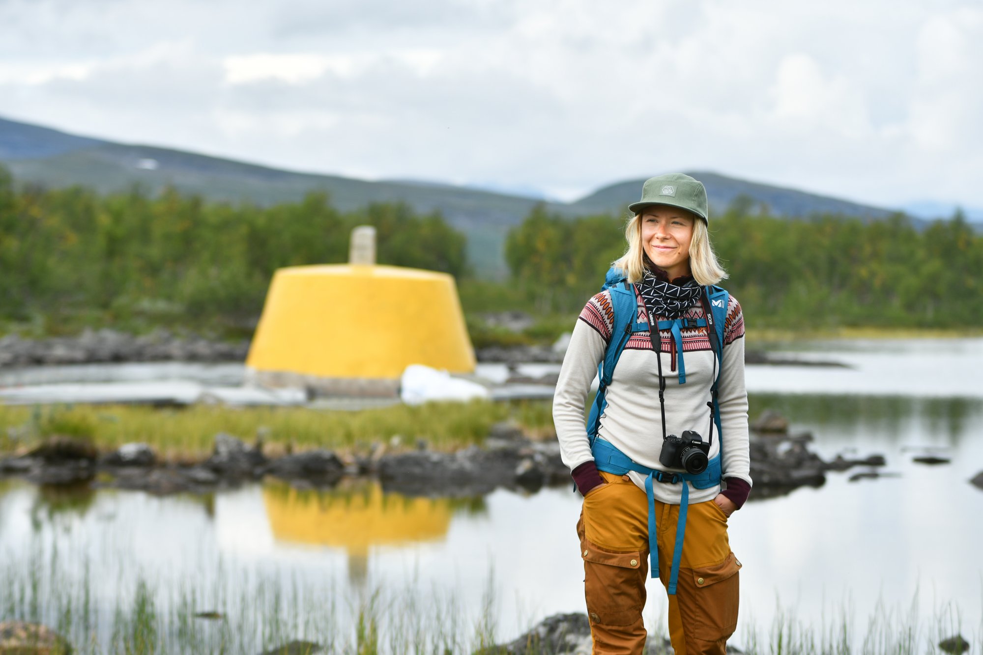 Nainen seisoo Kilpisjärvellä keltaisen Kolmen valtakunnan rajapyykin edessä vaellusvaatteissa kesällä