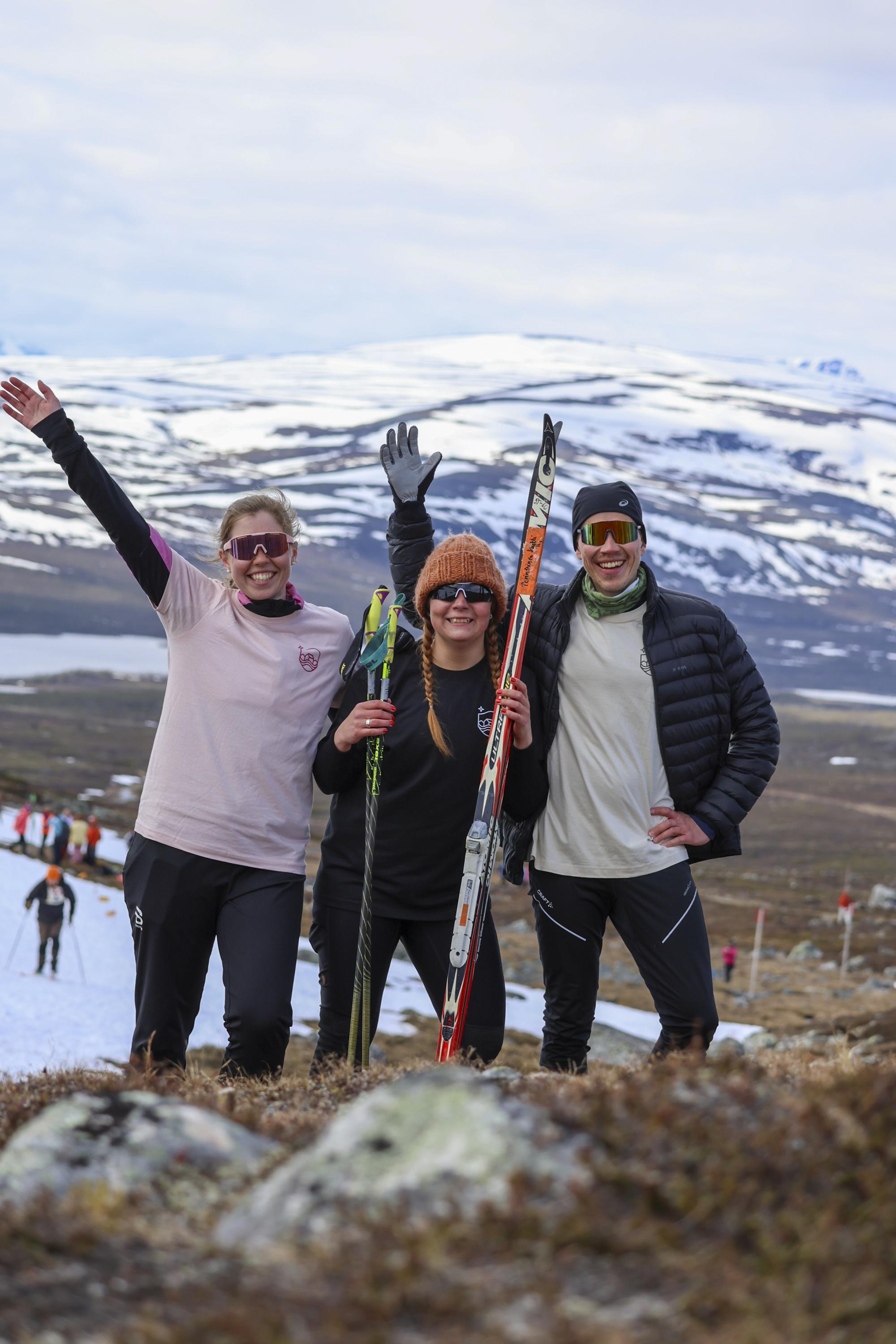 Three joyful Kilpisjärven Retkeilykeskus staff members have crossed the finish line at the Kilpisjärvi Midsummer Skiing event, smiling and raising their arms in celebration.