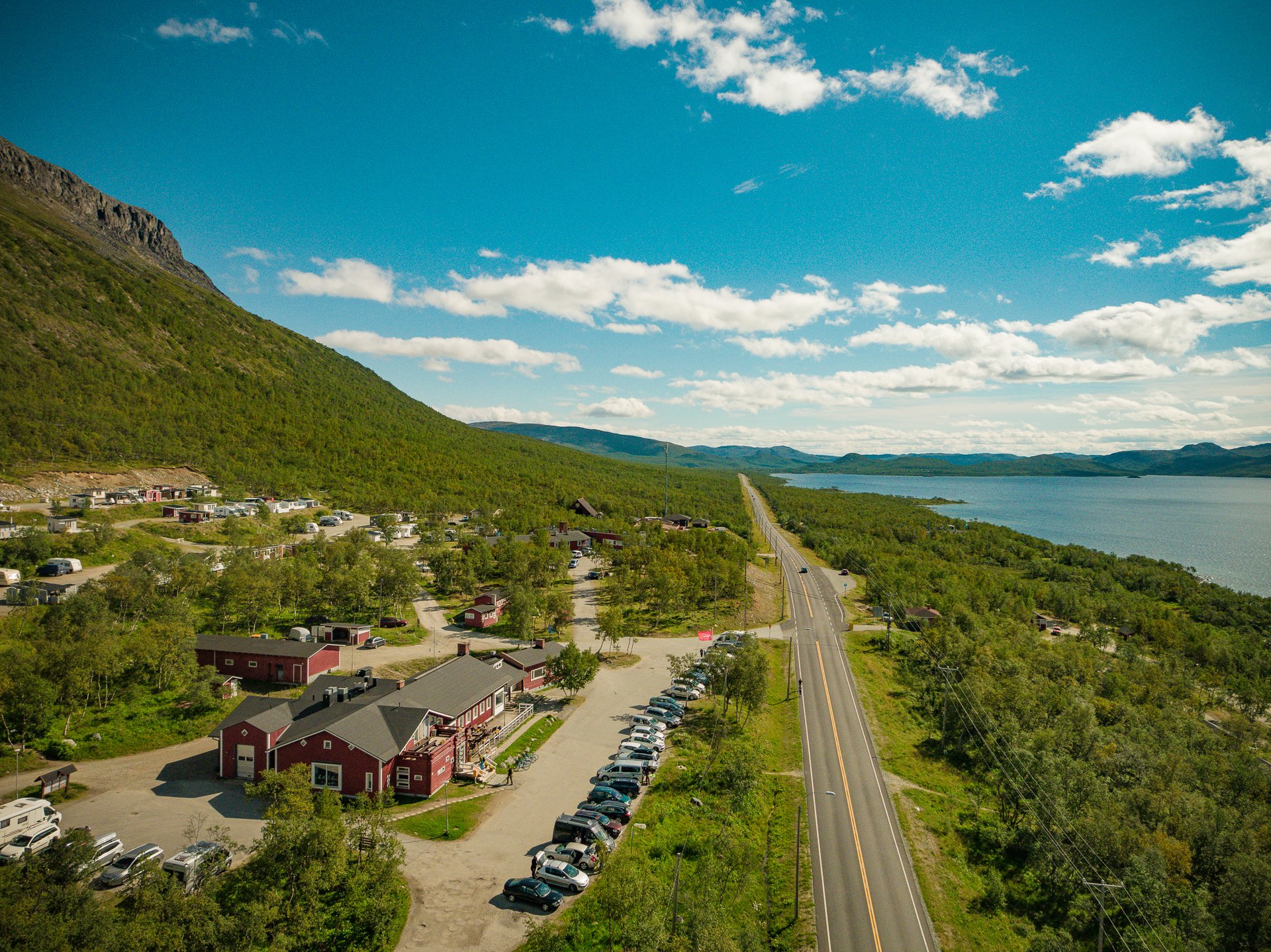 The camping site of Kilpisjärven Retkeilykeskus on a sunny summer day pictured from above. The sun is shining and you can see Lake Kilpisjärvi and Malla fell. 
