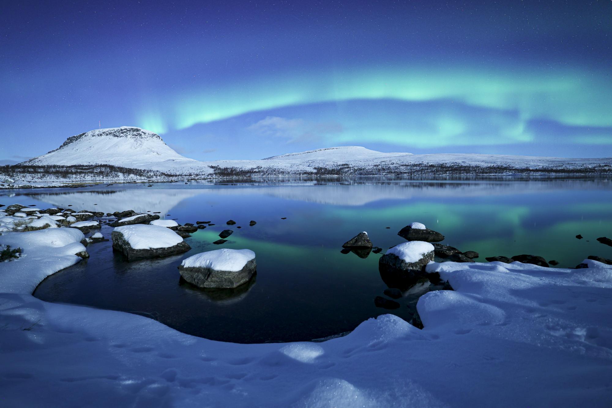 Spectacular Northern Lights above snow-covered Saana, with the still ice-free Lake Tsahkaljärvi in the foreground and snowy shores framing the scene.