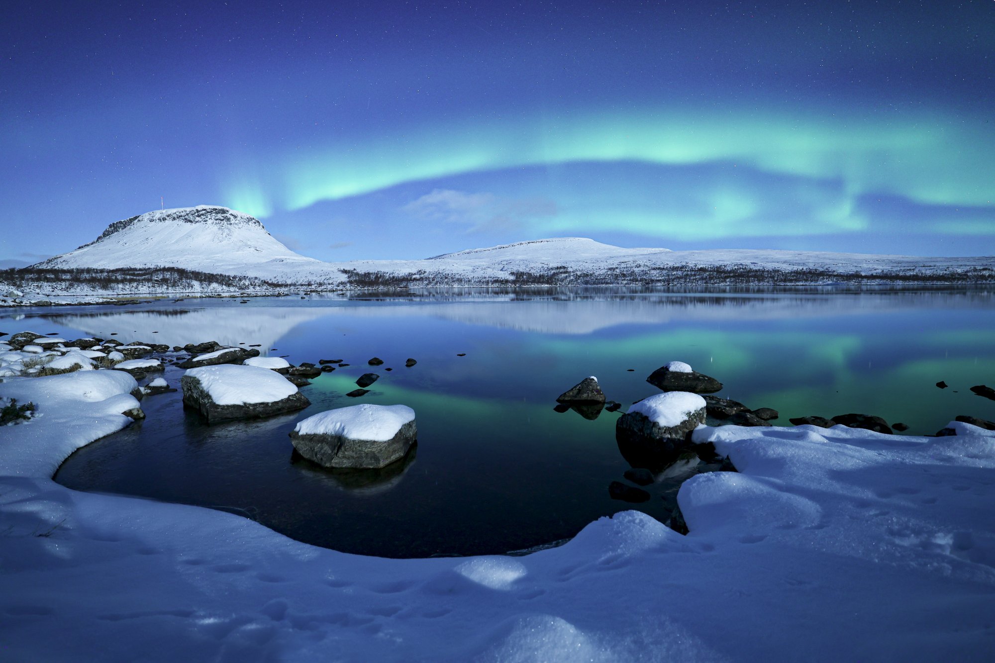 Spectacular Northern Lights above snow-covered Saana, with the still ice-free Lake Tsahkaljärvi in the foreground and snowy shores framing the scene.