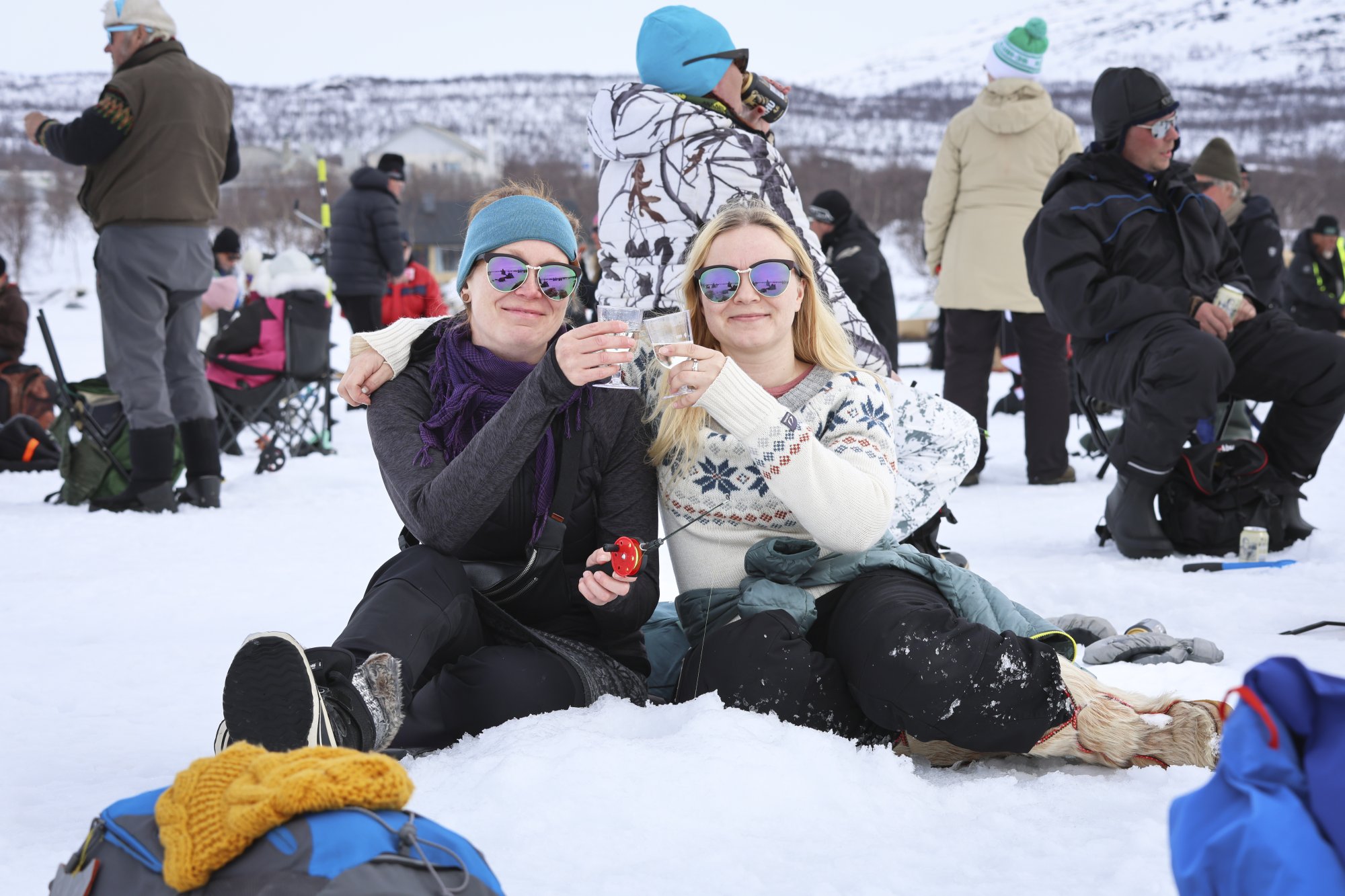 Two women clink glasses of sparkling wine while sitting on the ice of Lake Kilpisjärvi, wearing sunglasses. They are taking part in the Vain kaksi kalaa ice fishing event.