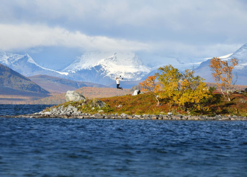 Lake Kilpisjärvi and a small island where a woman is jumping. Three trees are in autumn foliage and on the back grounfd you can see Norwegian mountains already covered in snow