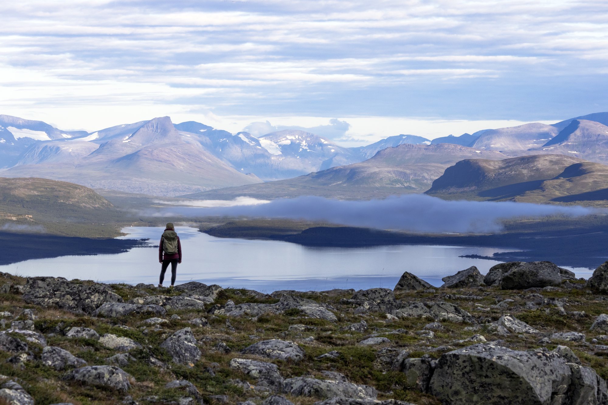 A woman is standing on a fell with lake and mountain landscape infront of her.