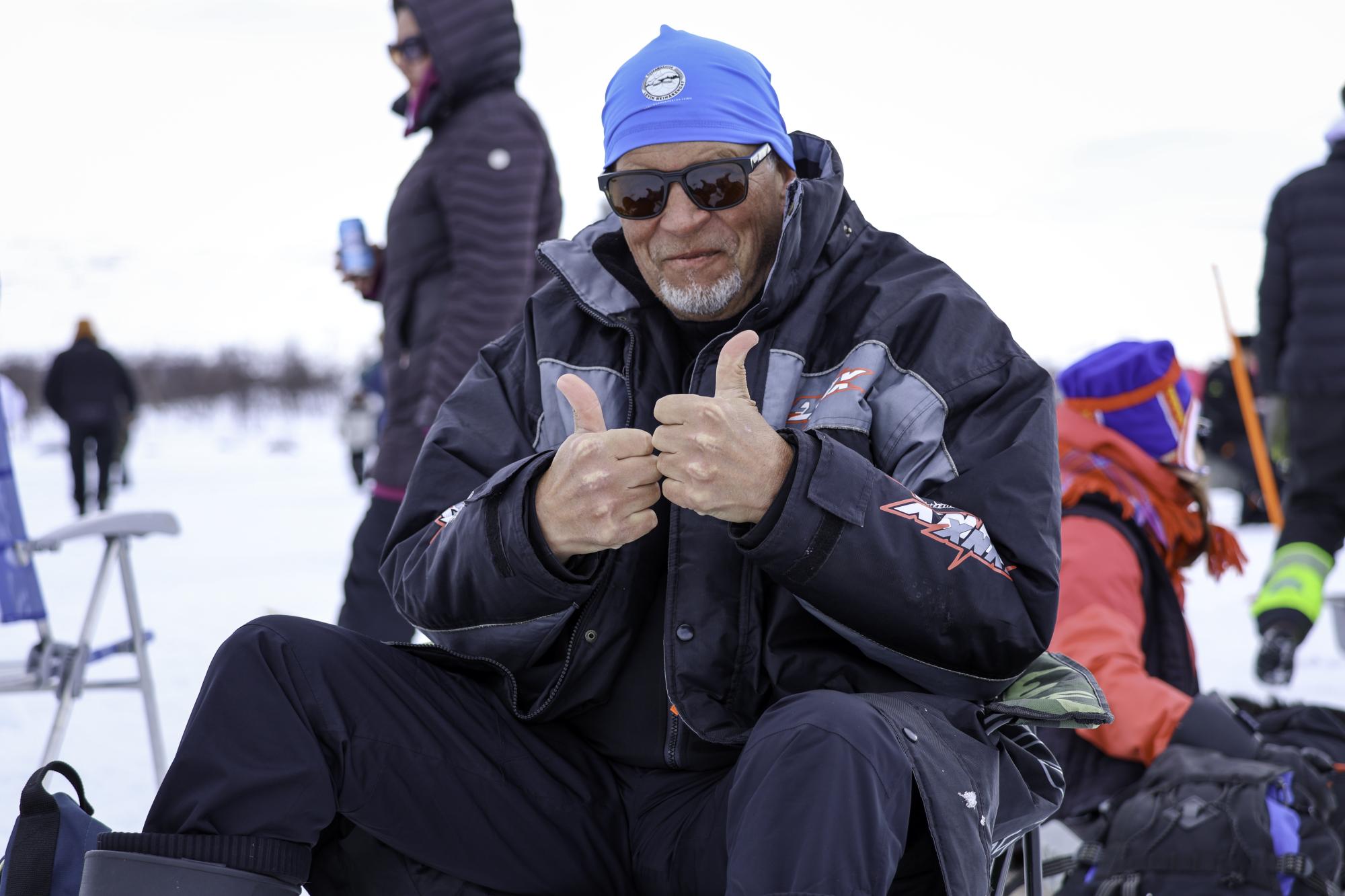 A joyful male ice fisher at the Vain kaksi kalaa event in Kilpisjärvi, smiling with both thumbs up.